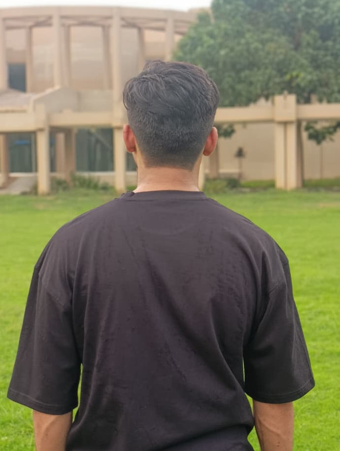 A man viewed from behind wearing a plain black t-shirt, standing on a green lawn with a modern beige building and trees in the background.