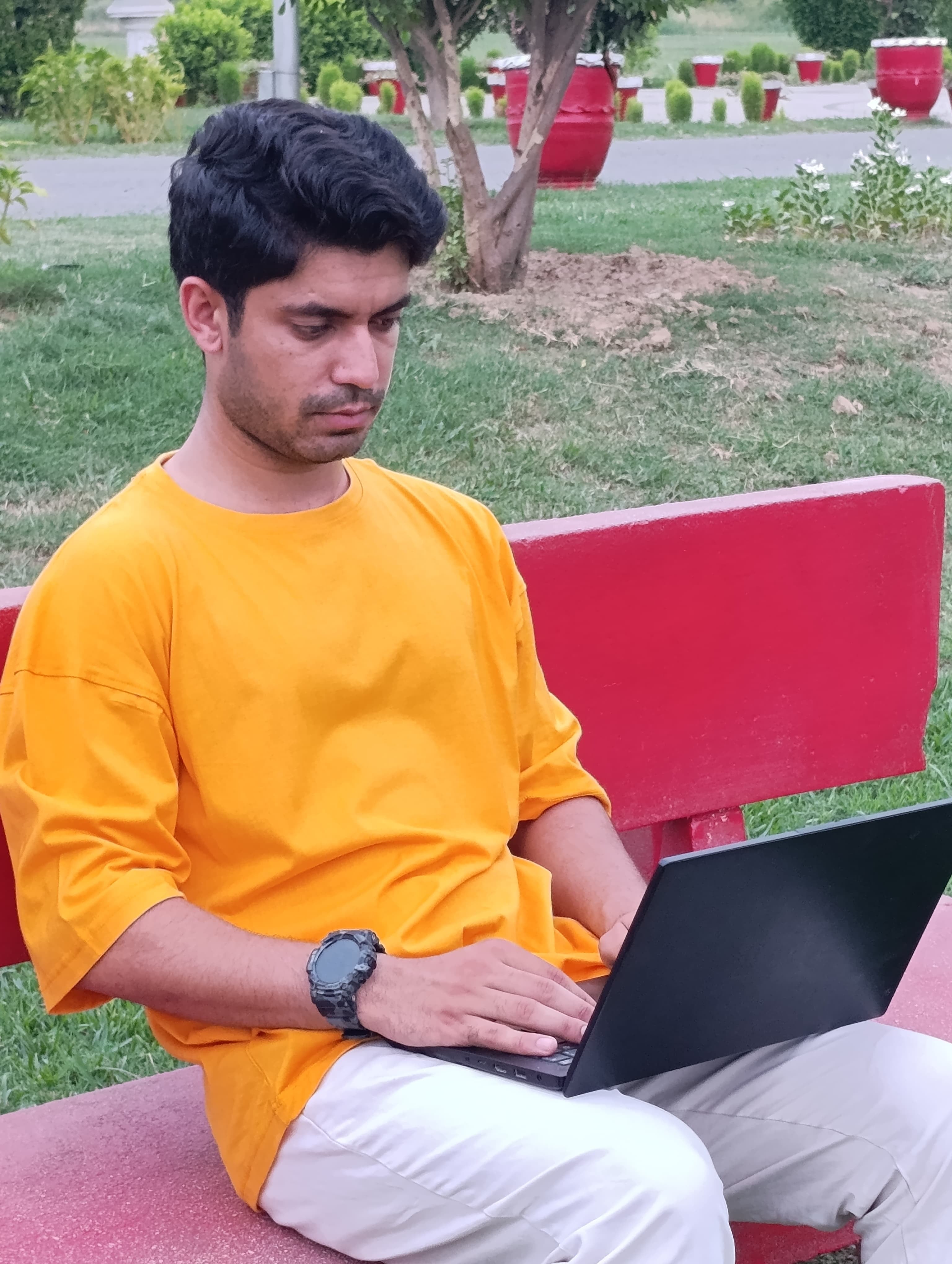 A man wearing a bright yellow t-shirt and off-white trousers sits on a red bench in a park, using a black laptop.