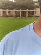 Close-up of a person wearing a light blue crewneck t-shirt outdoors, with a blurred background of green grass and a beige fence.