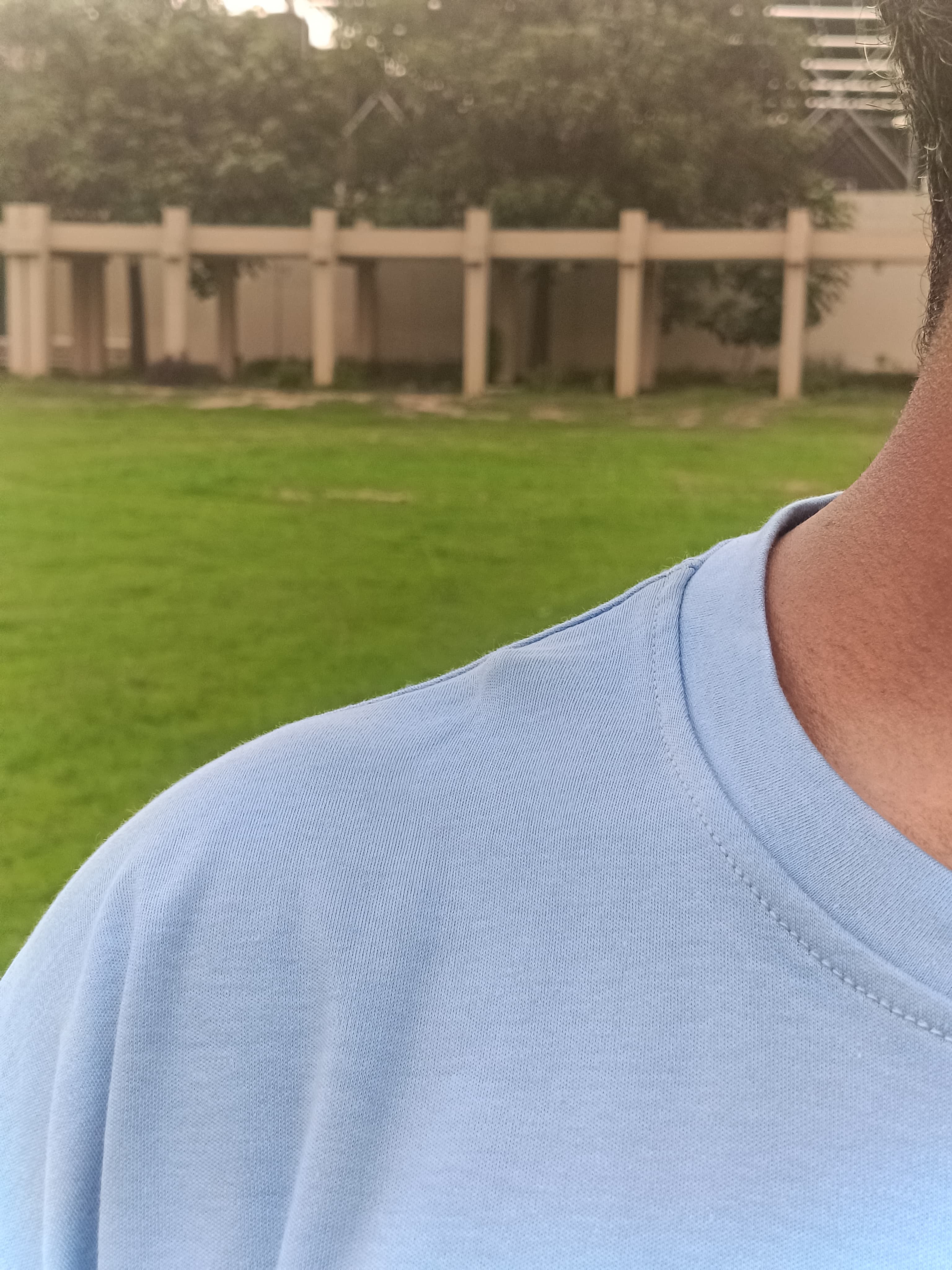 Close-up of a person wearing a light blue crewneck t-shirt outdoors, with a blurred background of green grass and a beige fence.