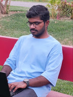 A man wearing a light blue t-shirt and glasses sits on a red bench in a park, using a black laptop.