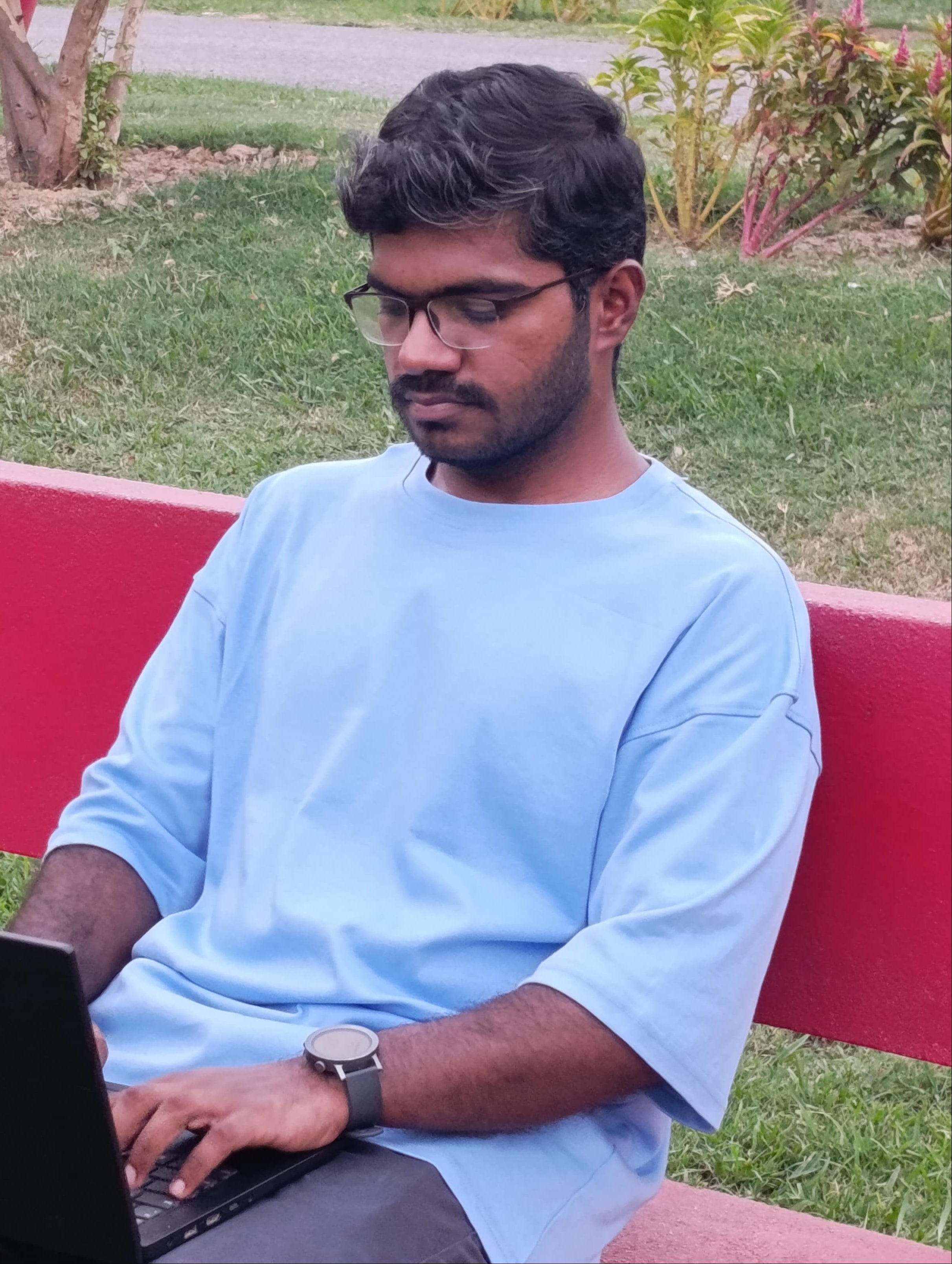 A man wearing a light blue t-shirt and glasses sits on a red bench in a park, using a black laptop.