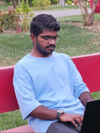 A man wearing a light blue t-shirt and glasses sits on a red bench in a park, using a black laptop.