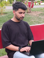 A man wearing a black t-shirt and light blue jeans sits on a red bench in a park, using a black laptop.