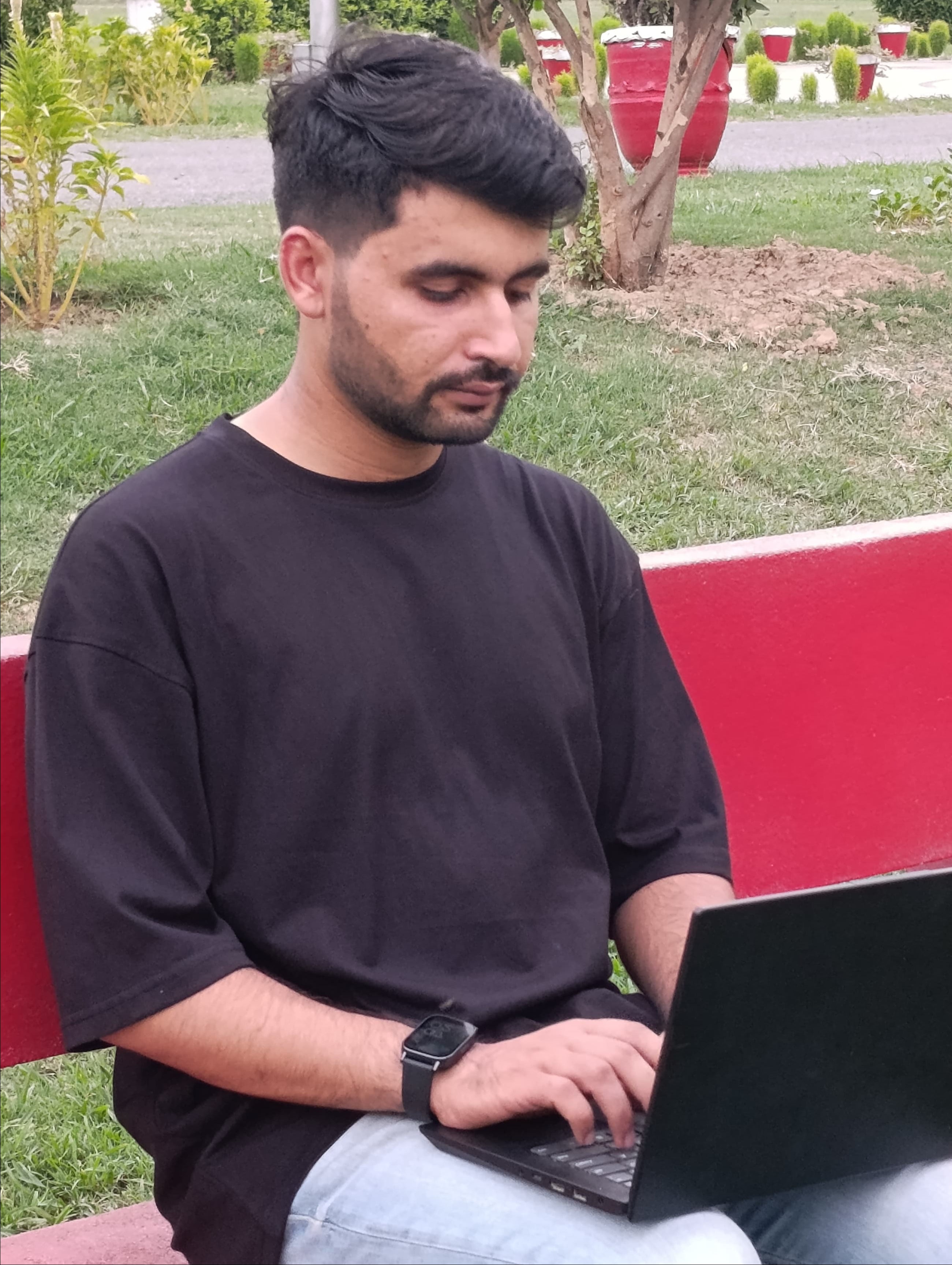 A man wearing a black t-shirt and light blue jeans sits on a red bench in a park, using a black laptop.