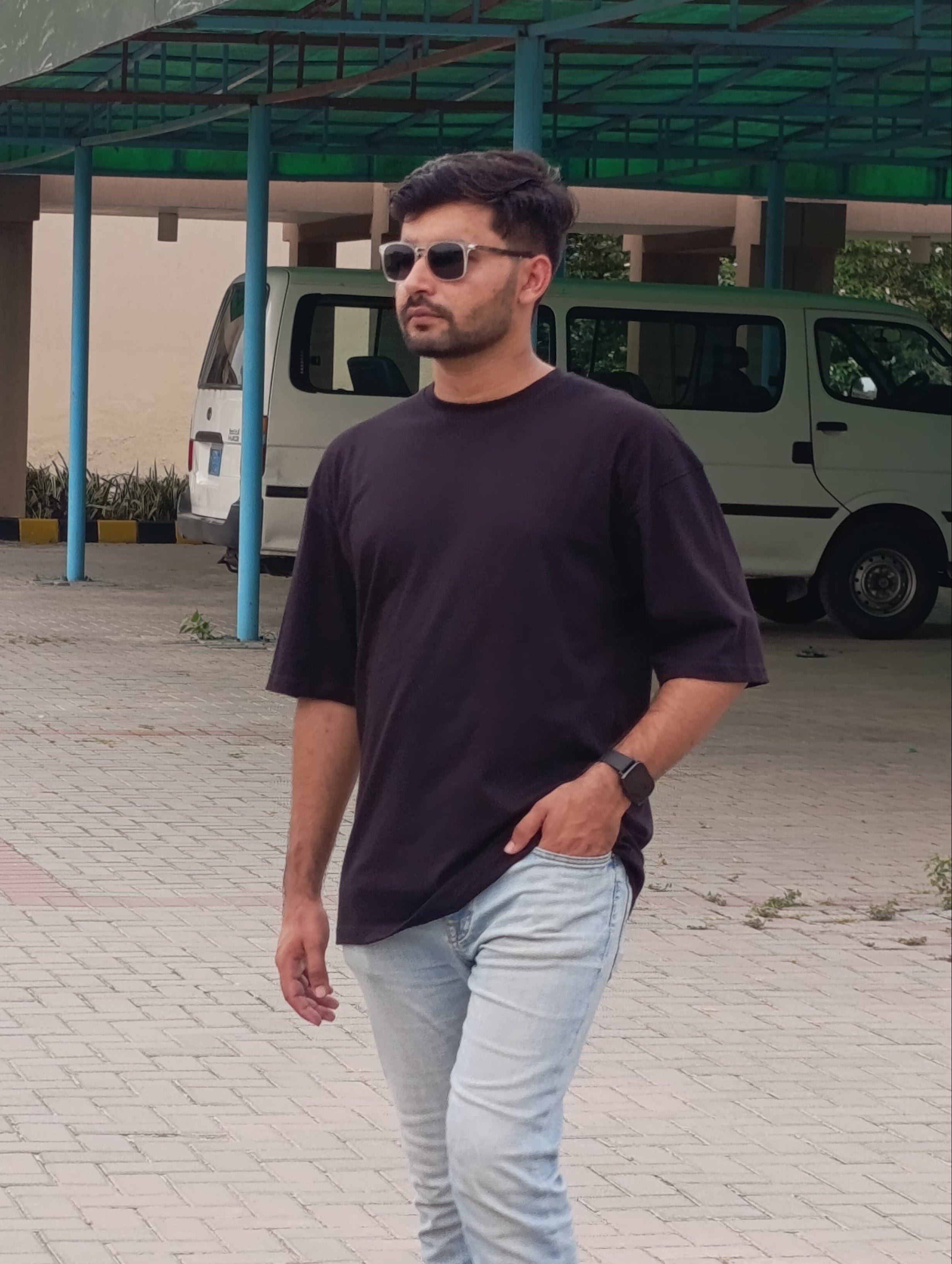 A man wearing a dark purple crewneck t-shirt and light blue jeans walking outdoors in front of a building with a green canopy and a white van.