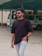 A man wearing a dark purple crewneck t-shirt and light blue jeans walking outdoors in front of a building with a green canopy and a white van.
